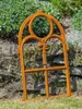 A rusty metal window frame leaning against a stone wall with greenery in the background. Produktbild-Vorschau