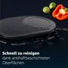 A close-up shot of a black oval raclette grill with some vegetables in the background. Produktbild-Vorschau
