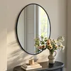 A round mirror with a black frame hangs on a wall above a dark console table. A vase of flowers and some books are on the table. Produktbild-Vorschau