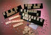 A collection of Rummikub tiles and racks arranged on a pinkish-grey surface, with scattered individual letter tiles. Náhľad obrázku produktu