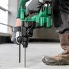A person wearing work gloves and boots is using a green and black cordless rotary hammer drill to drill into a concrete floor. Voir les images du produit