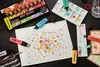 Art supplies arranged on a black surface, including markers, a palette with paint, and a paper with colorful dots. Anteprima dell’immagine prodotto