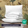 A stack of white bedding (a pillow and a folded duvet) is placed on a wooden surface in front of a nature-themed wallpaper with birds and foliage. A green OEKO-TEX logo is visible on the right side. Produktbild-Vorschau