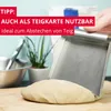 A hand is using a metal dough scraper to cut a piece of dough on a wooden surface. In the background, there are kitchen items and a potted plant. Produktbild-Vorschau