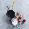 A top-down view of a small saucepan with a wooden handle, surrounded by plums, a jar of sugar, and sprigs of thyme on a textured gray surface. Náhled obrázku produktu