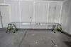 A long, silver plank supported by two green step ladders, set up on a concrete floor in what appears to be a garage or unfinished room. Produktbild-Vorschau