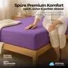 A person sitting on a bed with a purple fitted sheet. Produktbild-Vorschau