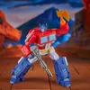 A red, blue, and white robot toy, identified as Optimus Prime, stands in a dynamic pose holding a gun and an axe. The background depicts a stylized, mountainous landscape with orange structures. Anteprima dell’immagine prodotto