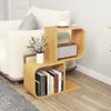 A modern S-shaped bamboo bookshelf is placed next to a sofa in a living room setting. The bookshelf holds books, a decorative vase with flowers, and a small speaker. A cup of coffee sits on top of the bookshelf. Podgląd zdjęcia produktu