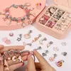 A person's hands are shown assembling a charm bracelet with various pink and silver charms, including unicorns, shells, and flowers. A pink jewelry box filled with more charms and beads is visible in the background. Produktbild-Vorschau