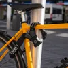 A yellow bicycle is locked to a silver pole with a black chain lock. The lock is attached to the frame of the bicycle and the pole. Produktbild-Vorschau