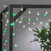 A string of LED party lights with white and green spherical bulbs is draped over a dark metal frame, with a blurred brick wall and greenery in the background. Náhled obrázku produktu