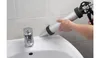 A person is using a caulk gun to apply sealant to the edge of a white sink next to a faucet. The background consists of light-colored tiled walls. Podgląd zdjęcia produktu