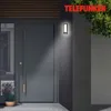 A modern dark gray front door with a long silver handle with wooden accents, illuminated by a wall-mounted LED light. A pot of lavender sits to the left of the door. Produktbild-Vorschau