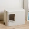 A modern, enclosed cat litter box with a clear plastic door, placed on a wooden floor next to a white wall and a bookshelf. Voir les images du produit