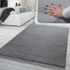 A soft, dark gray rug is shown in a living room setting with a white sofa and a close-up inset of a hand pressing into the rug to demonstrate its softness.