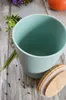 A turquoise ceramic storage jar with a wooden lid sits on a weathered wooden surface, surrounded by scattered peppercorns and a sprig of greenery. Náhľad obrázku produktu