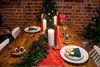 A festive Christmas table setting with a red runner, pine garland, candles, and place settings. Produktbild-Vorschau