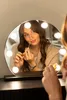 A woman applying makeup in front of a lighted vanity mirror. Náhled obrázku produktu