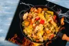 A frying pan filled with colorful vegetables is placed on a black surface with flames and water splashes, suggesting cooking. Produktbild-Vorschau