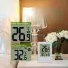 A digital thermometer showing indoor and pool temperatures, placed on a nightstand next to a lamp and flowers. Produktbild-Vorschau