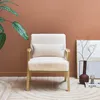 A fluffy white armchair is placed in a room with a brown wall and a patterned rug. A plant is on the left, and a vase with branches and a small sculpture are on the right. Produktbild-Vorschau