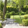 A black metal tripod stand is set up on a stone pathway in a garden with manicured bushes and trees. Náhled obrázku produktu