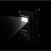 A black solar power bank with its flashlight on, attached to a black fabric holder, against a black background. Produktbild-Vorschau
