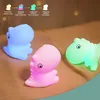 Three dinosaur-shaped LED night lights in blue, pink, and green are displayed on a textured surface. Voir les images du produit
