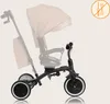 A product shot of a beige and black children's tricycle with a sun canopy. The tricycle is shown from a side-angle, with a blurred background. Podgląd zdjęcia produktu
