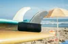 A black JBL speaker sits on a surfboard on a beach, with surfboards and a beach umbrella in the background. Voir les images du produit