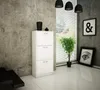 A white shoe cabinet with three drawers stands against a textured gray wall, next to a window with blinds and a potted plant. Artwork hangs on the wall. Produktbild-Vorschau