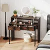 A vanity desk with makeup and accessories, set up in a bedroom next to a bed and a floor lamp. Náhled obrázku produktu