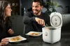 A man and a woman are sitting at a table, eating from plates. A rice cooker is on the table next to the man. The man is stirring the rice cooker with a wooden spoon. The woman is looking at the man and smiling. The scene is set in a modern kitchen. Podgląd zdjęcia produktu