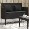 A black poly rattan garden bench with cushions is shown on a patio with a rug and a small coffee table. Náhled obrázku produktu