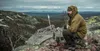 A man sits on a rocky outcrop in a mountainous landscape, wearing outdoor clothing. Náhled obrázku produktu