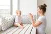 A mother and baby are sitting on a wooden bench by a window, with pillows behind them. The mother is clapping her hands and smiling at the baby, who is looking at her. Produktbild-Vorschau