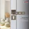 A close-up of a bronze-colored door handle set with a keyhole escutcheon, mounted on a white door. The background shows a blurred interior scene with furniture and decor. Produktbild-Vorschau