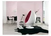 A modern living room with a pink wall, a white egg chair with red interior, a black rug, a stack of books, a table with chairs, and a bookshelf. Produktbild-Vorschau