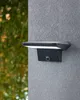A black outdoor solar lamp with a motion sensor and twilight sensor is mounted on a textured gray wall. Green foliage is visible in the blurred background. Náhled obrázku produktu