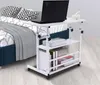 A white adjustable laptop table with shelves is placed next to a bed with pillows and a blanket. Náhled obrázku produktu