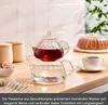 A glass teapot filled with red liquid and water, placed on a kitchen counter next to a cup of tea and a cake under a dome. Produktbild-Vorschau