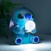 A blue Stitch figurine holding a glowing light bulb, sitting in a dimly lit room with a plant in the background. Produktbild-Vorschau