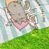 A close-up view of a Pusheen blanket with a cartoon cat sleeping on pillows, surrounded by gaming accessories and a box of donuts. The blanket is laid out on green grass. Produktbild-Vorschau