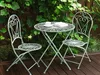 A green metal bistro set with a table and two chairs is set up on a wooden deck, surrounded by lush green foliage. Náhľad obrázku produktu