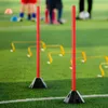 Two red training poles with black bases are set up on a green turf field, with yellow agility hurdles in the background and blurred figures of people. Náhled obrázku produktu