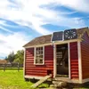 A solar-powered fan and solar panel are mounted on the roof of a red chicken coop in a grassy field under a blue sky with white clouds. Produktbild-Vorschau