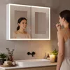 A woman brushing her teeth in front of a lit medicine cabinet above a sink. Podgląd zdjęcia produktu