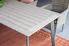 A close-up view of an outdoor dining table with a light teak Vironwood tabletop and gray metal legs, placed on an orange rug on a tiled patio. A wicker chair is visible in the background. Produktbild-Vorschau