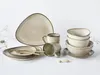 A collection of ceramic tableware, including plates, bowls, and mugs, arranged on a white surface with some dried plant sprigs. Produktbild-Vorschau